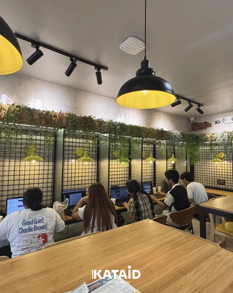 Five students working on laptops along a wall desk under black and yellow pendant lamps at Kataid.