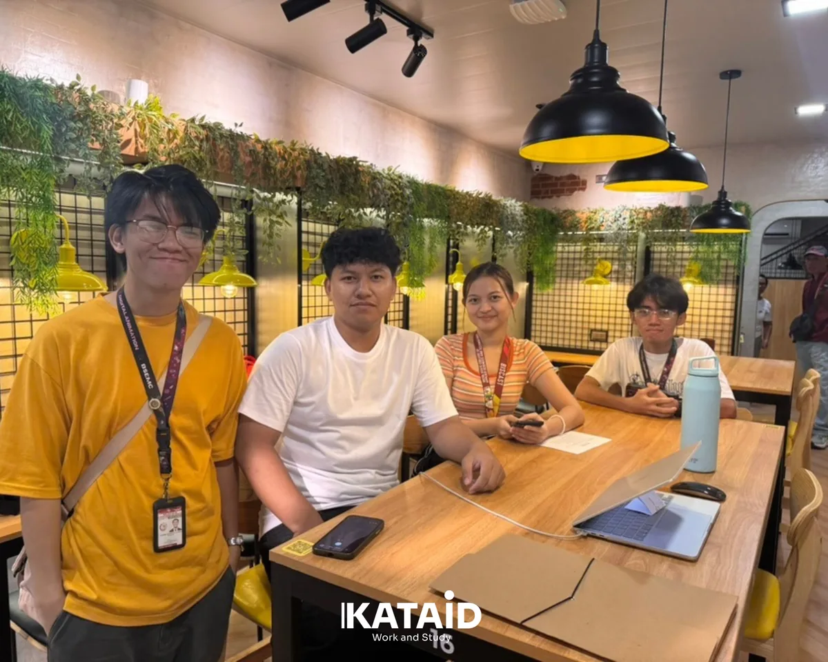 Four BISCAST students at a wooden table inside Kataid, under black pendant lamps and a wall of hanging plants.