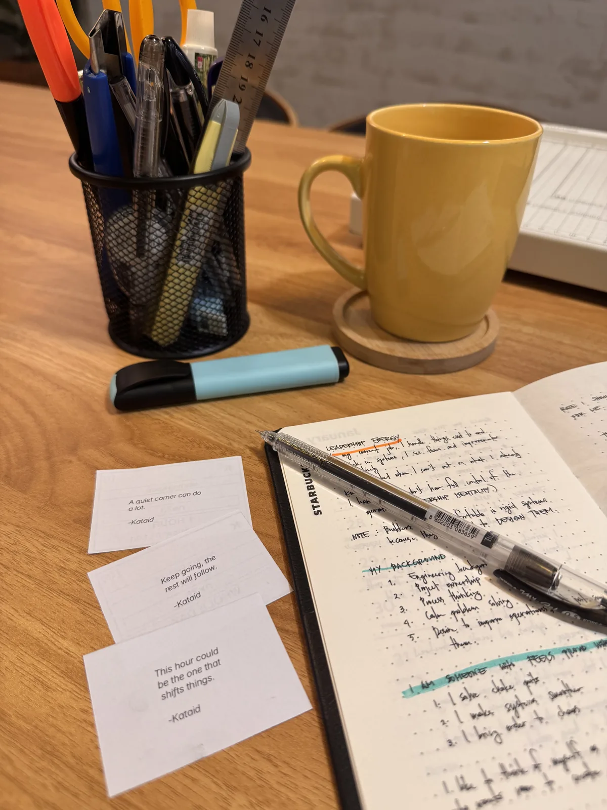 A desk at Kataid with a yellow mug, a cup of pens, an open notebook, and three Kataid WiFi cards.